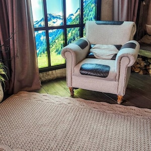 Op de afbeelding: Een lichtbruin gebreid tapijt met een golvende rand, geplaatst voor een groot raam met uitzicht op besneeuwde bergen en een stoel met een bruin lederen kussen.