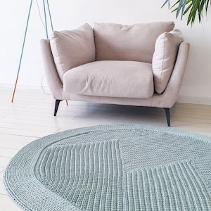 Op de afbeelding: Een lichtblauw ovaal gebreid vloerkleed met een textuurpatroon. Het vloerkleed ligt op een lichte houten vloer in een woonkamer.