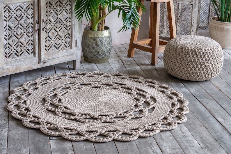 May include: A beige crocheted round rug with a delicate lace pattern. The rug is placed on a wooden floor with a wooden stool and a potted plant in the background.