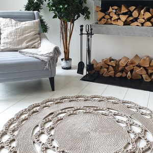 May include: A beige crocheted round rug with a delicate lace pattern. The rug is placed in a living room setting with a fireplace and a potted plant.