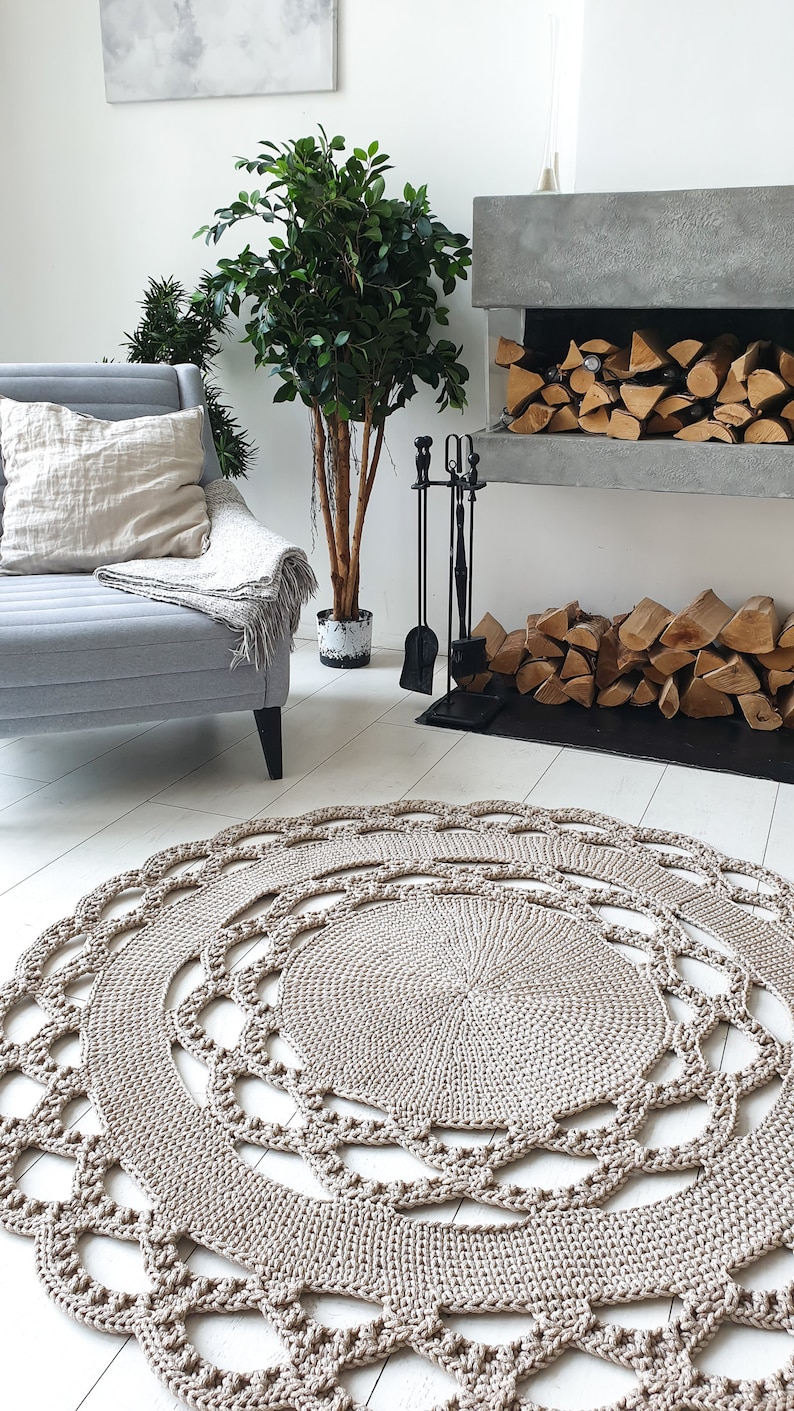 May include: A beige crocheted round rug with a delicate lace pattern. The rug is placed in a living room setting with a fireplace and a potted plant.