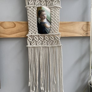 May include: A macrame photo frame in natural beige, featuring a rectangular opening holding a portrait of a child. The frame is suspended from a wooden branch and includes a fringe of knotted cords.