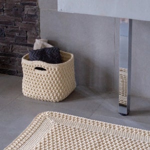 May include: A cream-colored crocheted bath mat with a textured design. The mat is placed on a tiled floor in a bathroom setting.