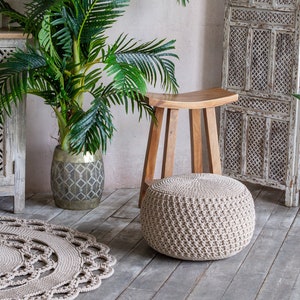 May include: A beige crocheted pouf ottoman sits on a wooden floor in front of a wooden stool. A potted palm tree and a white wooden screen are in the background.