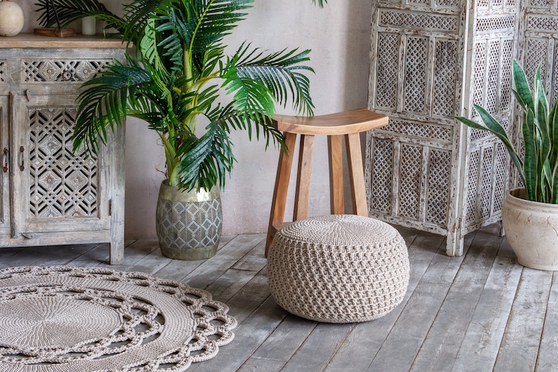 May include: A beige crocheted pouf ottoman sits on a wooden floor in front of a wooden stool. A potted palm tree and a white wooden screen are in the background.