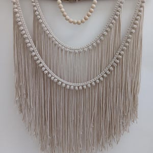 May include: A beige macrame wall hanging suspended from a natural wood branch. The design includes two curved, fringed sections and a beaded accent. The macrame is made of a soft, woven material, creating a bohemian aesthetic.