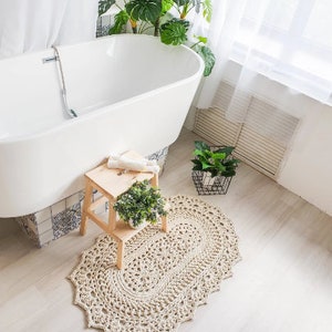 以下が含まれることがあります： 植物のあるバスルームに、白いバスタブ、木製のスツール、かぎ針編みのラグが置かれています
