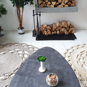 May include: Two beige crocheted round rugs with a decorative pattern, a gray oval coffee table with a green plant and a glass bowl of decorative items, a gray couch with a throw blanket, a fireplace with a black metal grate and a stack of logs, and a potted plant in the background.