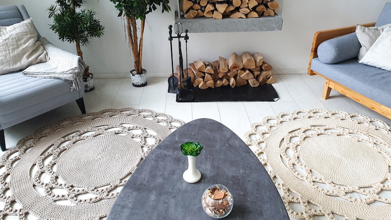 May include: Two beige crocheted round rugs with a decorative pattern, a gray oval coffee table with a green plant and a glass bowl of decorative items, a gray couch with a throw blanket, a fireplace with a black metal grate and a stack of logs, and a potted plant in the background.