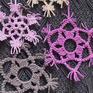 May include: Close-up of a wall hanging with multiple crocheted snowflakes in shades of gray, pink, and beige. The snowflakes are hanging from a gray macrame background.