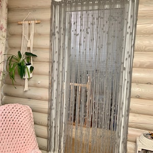 May include: A gray macrame curtain hangs in a doorway, with a scalloped top edge and knotted details. The curtain is suspended from a wooden rod. A macrame plant hanger and a pink knitted blanket are visible in the background.