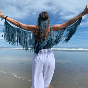 May include: A woman wearing a blue crocheted top with fringe and a white crocheted headpiece. She is standing on a beach with her arms raised.