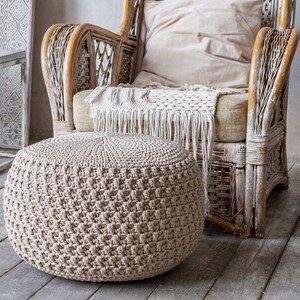 May include: A beige crocheted ottoman with a woven texture sits in front of a white wicker armchair with a beige cushion and a white throw blanket. The chair and ottoman are in a room with a light wood floor.