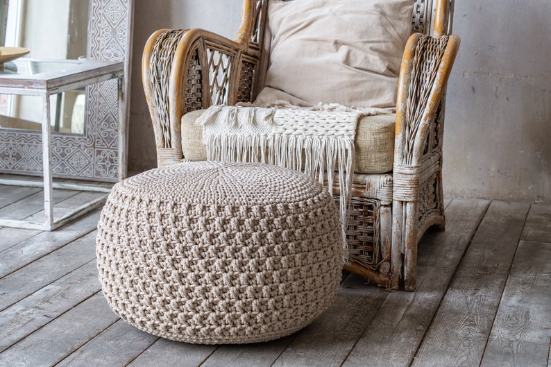 May include: A beige crocheted ottoman with a woven texture sits in front of a white wicker armchair with a beige cushion and a white throw blanket. The chair and ottoman are in a room with a light wood floor.