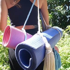 以下が含まれることがあります： 黒色の服を着た人が、白と青の編み紐で結ばれた、巻かれたヨガマットを2つ持っています。紐には白と青の房が付いています。