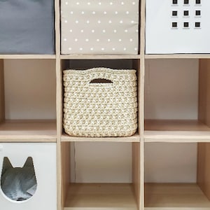 Pode incluir: Uma estante de cubos branca com seis compartimentos. Os três compartimentos superiores estão cheios de caixas de armazenamento. Os três compartimentos inferiores estão vazios. As caixas de armazenamento são uma caixa de tecido cinza, uma caixa de tecido bege com bolinhas brancas e uma cesta de crochê bege.
