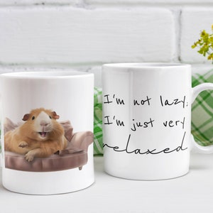 May include: Two white ceramic mugs with black text. One mug features a cartoon guinea pig relaxing on a couch with the text "I'm not lazy, I'm just very relaxed". The other mug has the same text but without the guinea pig.