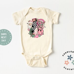 K&ouml;nnte beinhalten: Ein cremefarbener Baby-Strampler mit einem pinkfarbenen und gr&uuml;nen Weihnachtsmotiv. Das Motiv zeigt einen Weihnachtsmann, einen Weihnachtsbaum, einen Regenbogen, ein Karomuster und ein Smiley.