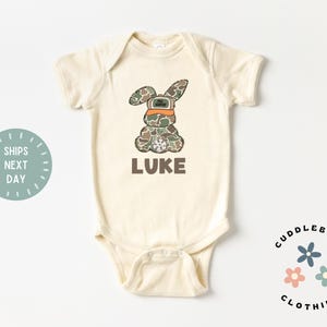 May include: A cream-colored baby onesie with a camouflage bunny wearing an orange hat and the name "LUKE" printed below. The onesie is from Cuddlebugs Clothing.