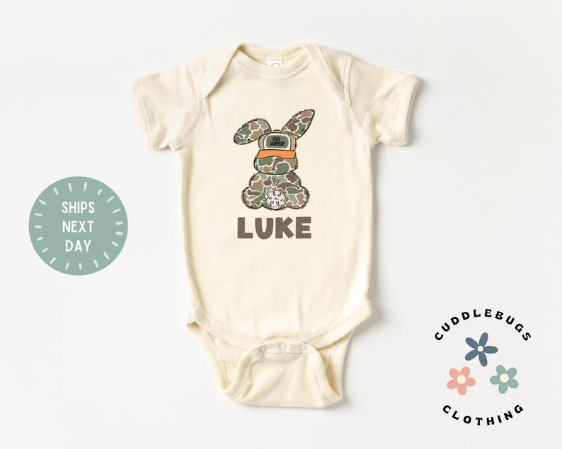 May include: A cream-colored baby onesie with a camouflage bunny wearing an orange hat and the name "LUKE" printed below. The onesie is from Cuddlebugs Clothing.