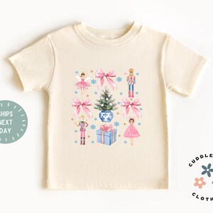 May include: A cream colored t-shirt with a Christmas themed design featuring a ballerina, a nutcracker, a Christmas tree, and a gift box. The design is in pink, blue, and green.