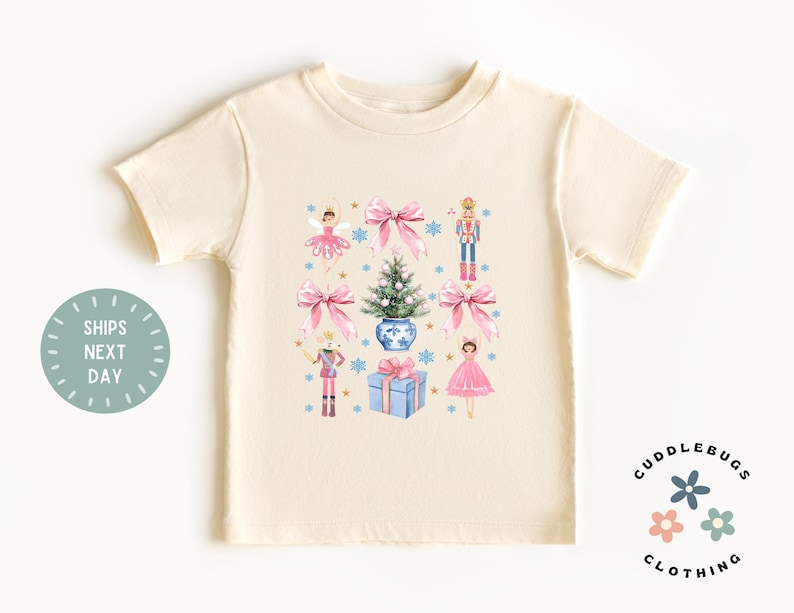 May include: A cream colored t-shirt with a Christmas themed design featuring a ballerina, a nutcracker, a Christmas tree, and a gift box. The design is in pink, blue, and green.