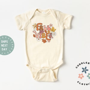 May include: Cream-colored baby bodysuit with the words "Fall Vibes" in a retro font, surrounded by fall-themed floral graphics in shades of orange, brown, and pink. The bodysuit is a one-piece garment, perfect for infants.