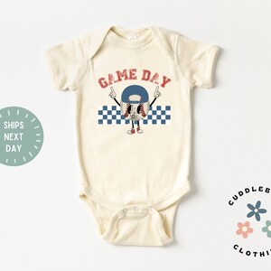 May include: Cream-colored baby bodysuit with a baseball-themed graphic. The graphic features a baseball character wearing a blue cap and red shoes, with the words "GAME DAY" in red above it. A blue and white checkered pattern is below the baseball character.