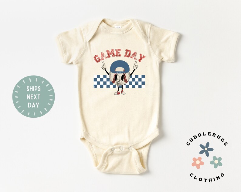 May include: Cream-colored baby bodysuit with a baseball-themed graphic. The graphic features a baseball character wearing a blue cap and red shoes, with the words "GAME DAY" in red above it. A blue and white checkered pattern is below the baseball character.