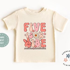 May include: A light beige t-shirt with a pink and white graphic that says "Five is a Vibe" with a peace sign and flowers.