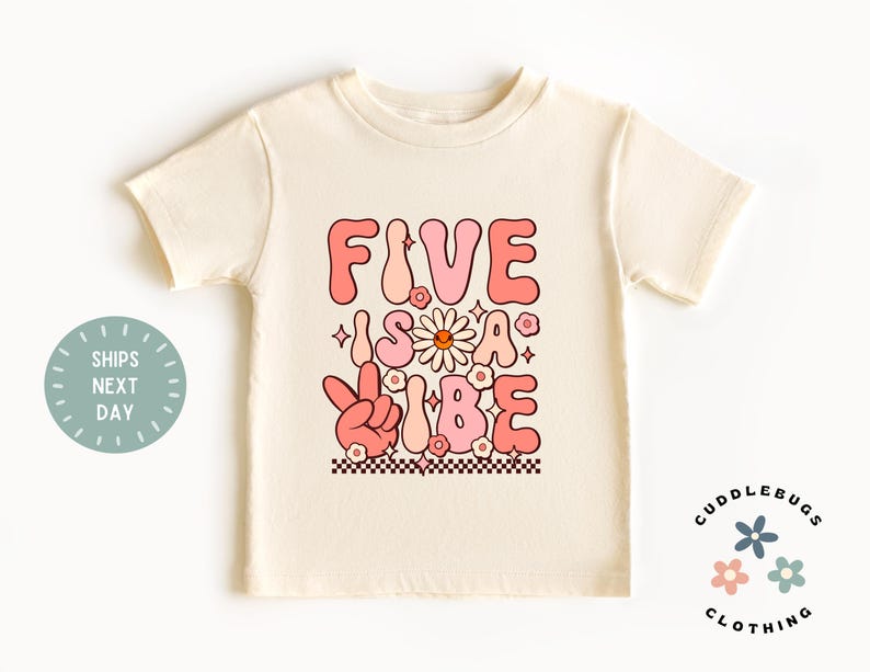 May include: A light beige t-shirt with a pink and white graphic that says "Five is a Vibe" with a peace sign and flowers.