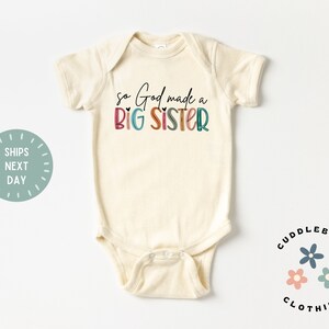 May include: A cream-colored baby onesie with the text "so God made a BIG SISTER" in colorful letters. The text is in a rainbow color scheme with a heart symbol between "God" and "made".