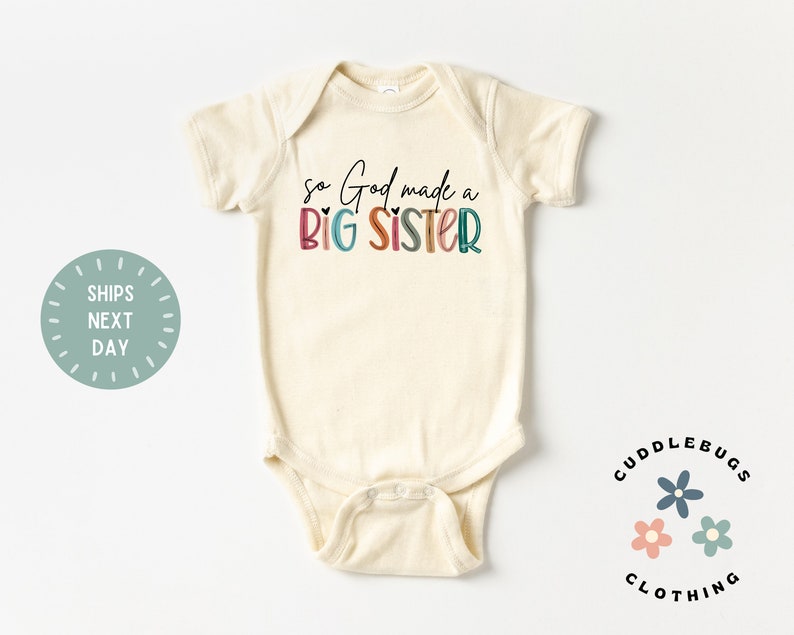 May include: A cream-colored baby onesie with the text "so God made a BIG SISTER" in colorful letters. The text is in a rainbow color scheme with a heart symbol between "God" and "made".