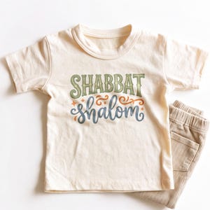 May include: A cream-colored short-sleeve t-shirt featuring the text SHABBAT in green and shalom in blue, accompanied by orange decorative swirls and small star accents. The shirt is paired with a pair of light brown corduroy pants.