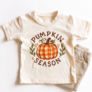 May include: A cream-colored short-sleeve t-shirt featuring a central graphic of an orange and white gingham pumpkin. The text "PUMPKIN SEASON" is printed in brown letters around the pumpkin, accented by small green leaves and orange hearts.