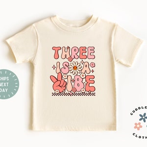 Pu&ograve; includere: Una maglietta beige per bambini piccoli con una grafica rosa e bianca che dice "Three is a vibe" con fiori e un segno di pace. La maglietta &egrave; di Cuddlebugs Clothing.