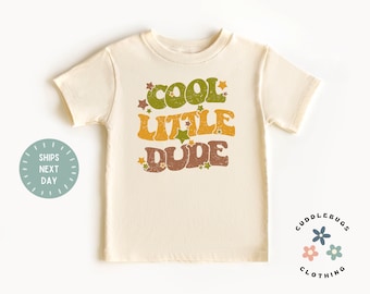 May include: A light beige short-sleeved t-shirt with the text "COOL LITTLE DUDE" in a retro font with stars. The text is in shades of green, yellow, and brown.