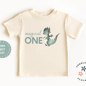 May include: Cream-colored toddler t-shirt with a teal dragon illustration and the words "magical ONE." The shirt has short sleeves and a round neckline. A circular graphic on the left says "SHIPS NEXT DAY." The Cuddlebugs Clothing logo is on the right.
