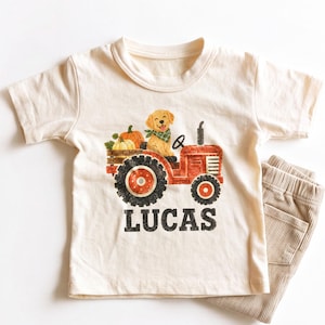 May include: A cream-colored short-sleeve t-shirt featuring a graphic of a golden retriever puppy wearing a green plaid bandana, sitting on a red tractor with pumpkins. The name LUCAS is printed in black capital letters below the tractor.