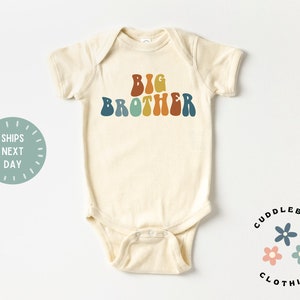 May include: A cream-colored baby onesie with the text "BIG BROTHER" in rainbow-colored letters. The onesie is from the brand "CUDDLEBUGS CLOTHING".
