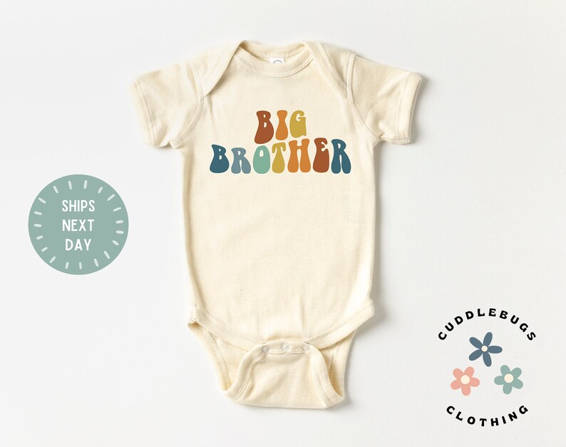 May include: A cream-colored baby onesie with the text "BIG BROTHER" in rainbow-colored letters. The onesie is from the brand "CUDDLEBUGS CLOTHING".