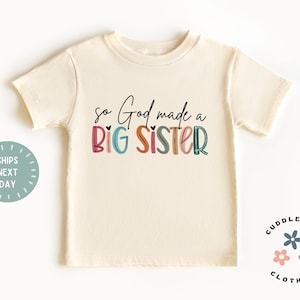 May include: A cream colored t-shirt with the text "so God made a BIG SISTER" in colorful letters. The text is in a playful font with hearts and a rainbow color scheme.