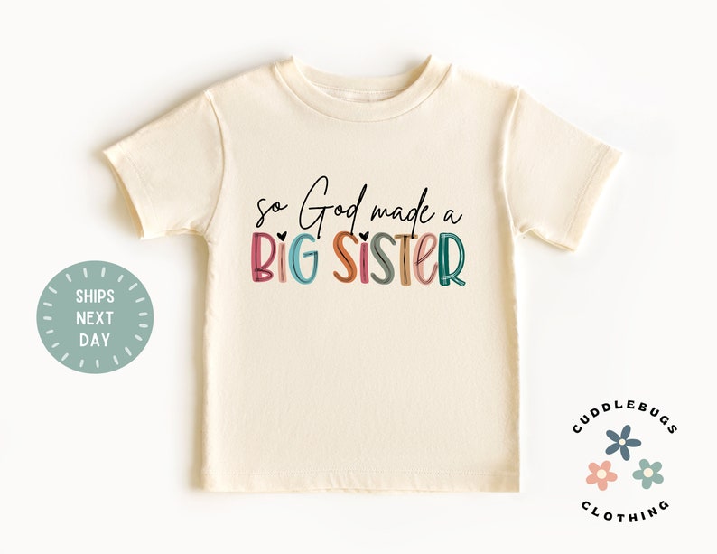 May include: A cream colored t-shirt with the text "so God made a BIG SISTER" in colorful letters. The text is in a playful font with hearts and a rainbow color scheme.