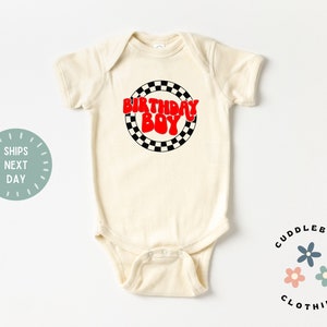 K&ouml;nnte beinhalten: Ein cremefarbener Baby-Strampler mit einem roten und schwarzen karierten Kreisdesign. Der Text "Birthday Boy" ist in roten Buchstaben innerhalb des Kreises geschrieben.