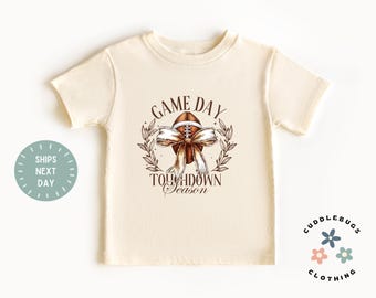 May include: Cream-colored t-shirt featuring a football graphic with a bow and the words "Game Day Touchdown Season." The design is surrounded by a wreath. The shirt has short sleeves and a round neckline.