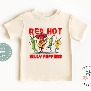 May include: Cream-colored toddler t-shirt with a playful graphic. The design features dancing chili peppers, one playing a guitar, with the words "RED HOT" and "SILLY PEPPERS" in red. The shirt has short sleeves and a round neckline.