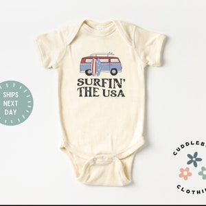 May include: A cream-colored baby onesie with a vintage-style illustration of a blue and red van with surfboards on top. The text "Surfin' The USA" is printed in brown below the van.