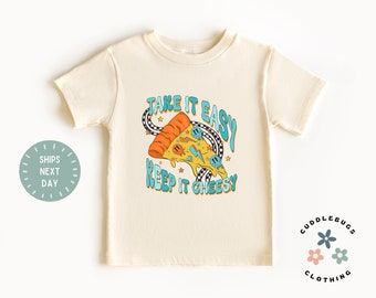 May include: A beige short-sleeved t-shirt with a graphic of a pizza slice with the text "Take it easy keep it cheesy" in blue and orange lettering.