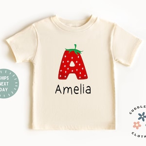 May include: A cream colored short-sleeved t-shirt with a red strawberry shaped letter A and the name Amelia printed below. The shirt has a small logo in the bottom right corner that says "Cuddlebugs Clothing" with three small flowers.