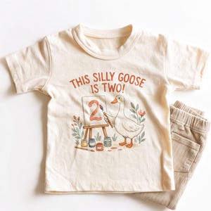 May include: A cream-colored short-sleeve t-shirt featuring a graphic of a goose painting the number two on an easel. The text above the illustration reads THIS SILLY GOOSE IS TWO! The shirt is paired with light tan corduroy pants visible on the side.